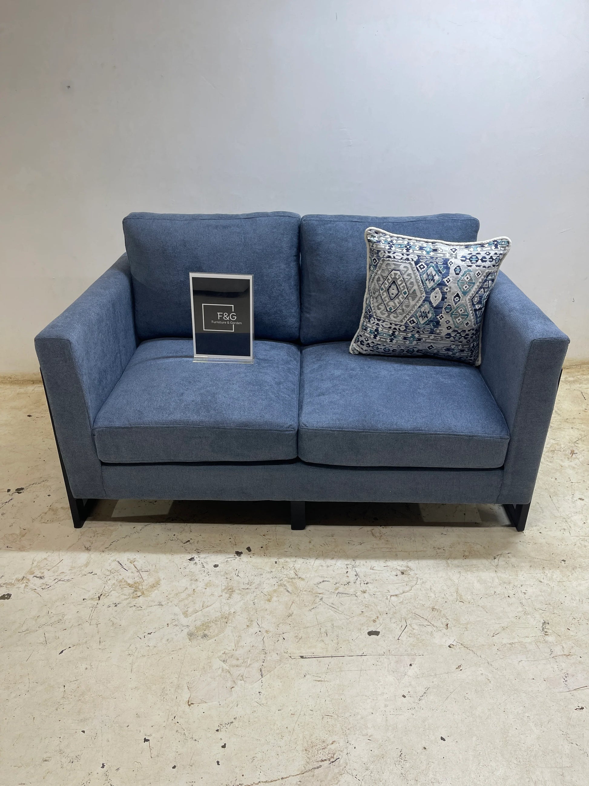 Blue fabric loveseat sofa with black legs, decorative patterned pillow, and F&G Furniture & Garden sign