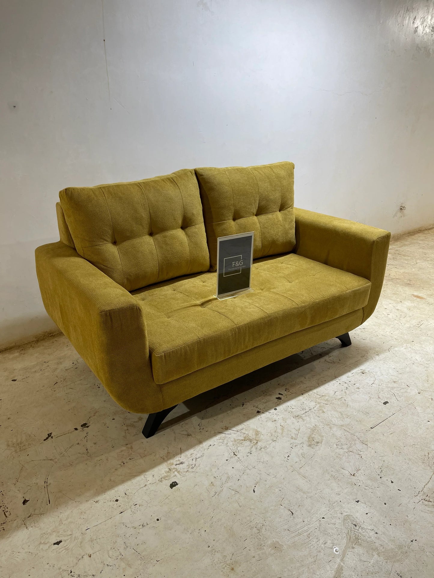 Mustard yellow modern two-seater sofa with tufted cushions on a concrete floor against a plain white wall