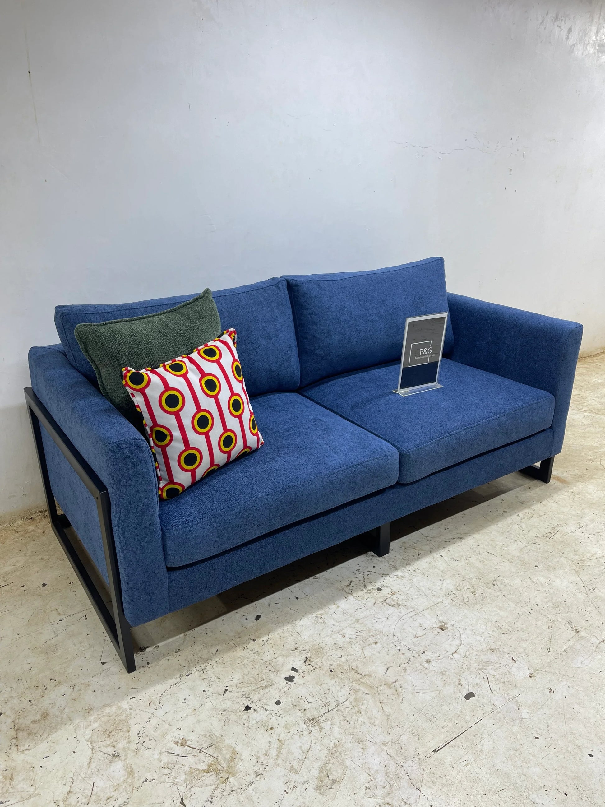 Blue fabric modern sofa with metal frame, featuring green and patterned decorative pillows on concrete floor