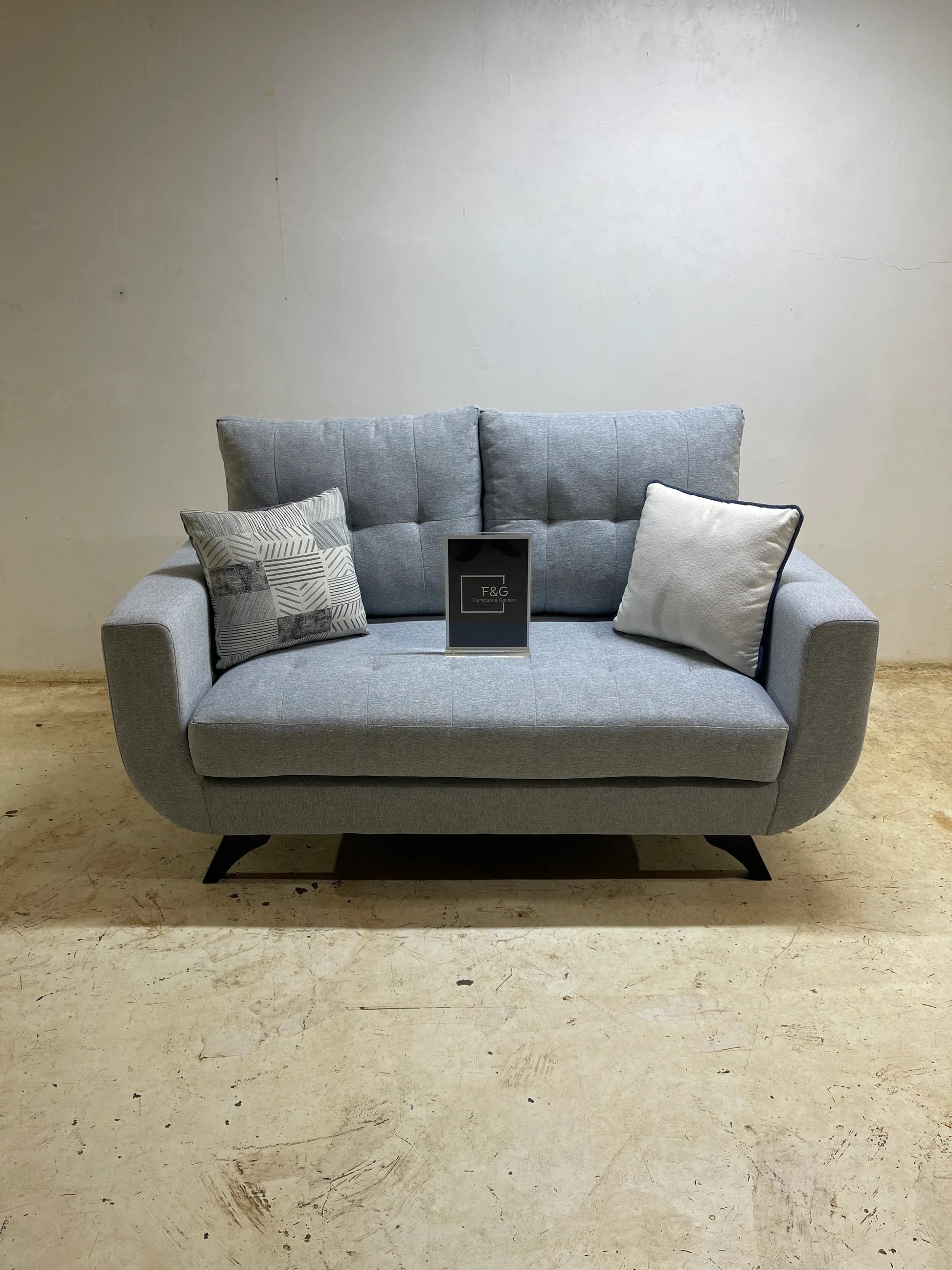 Gray modern loveseat sofa with two decorative pillows on rustic floor against a plain wall