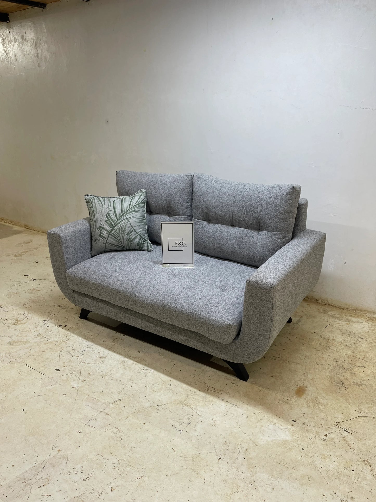 Modern gray fabric loveseat sofa with tufted cushions and green leaf patterned pillow on a light floor
