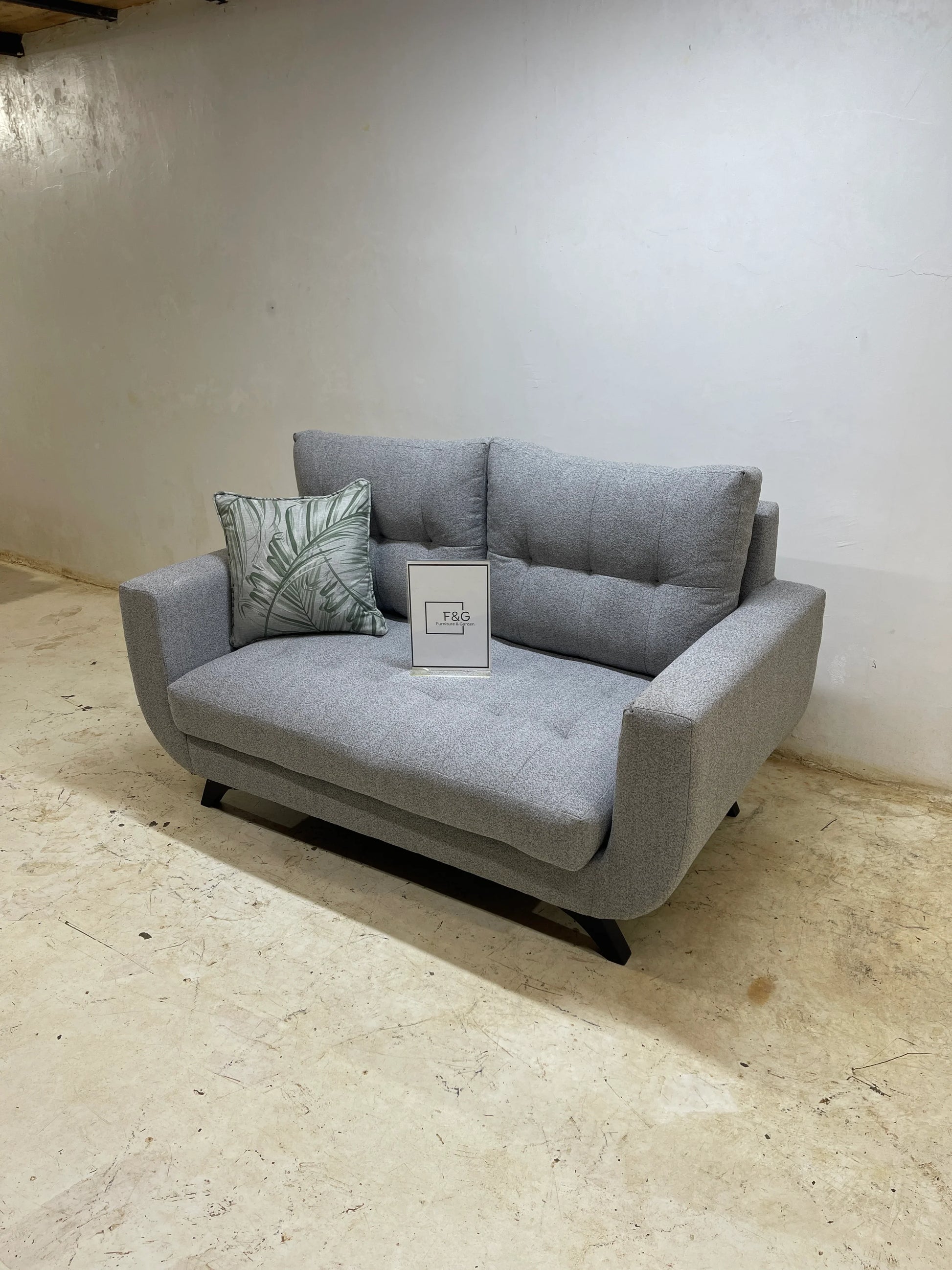Modern gray fabric loveseat sofa with tufted cushions and green leaf patterned pillow on a light floor