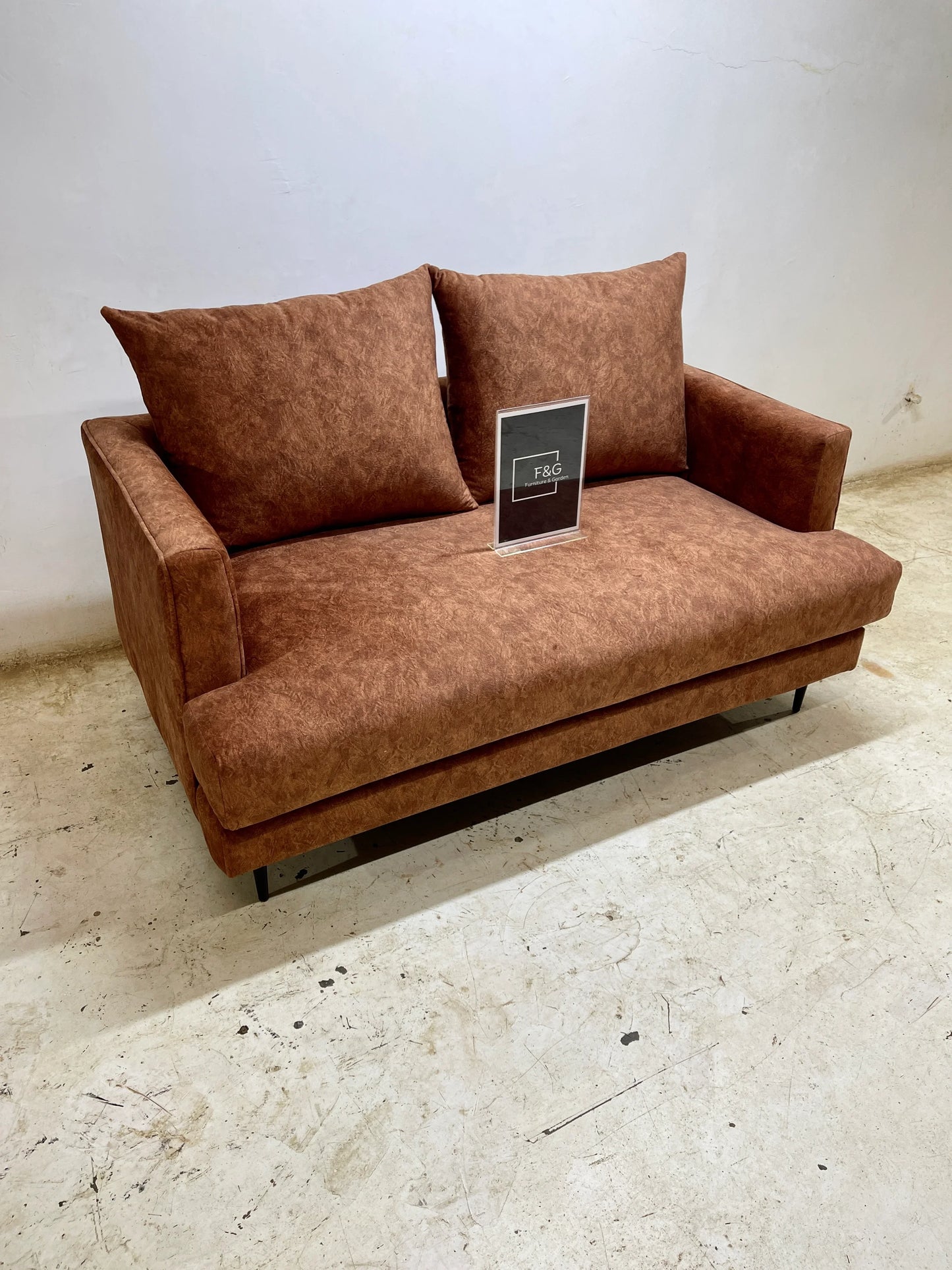 Brown fabric loveseat with two large cushions and black legs on a concrete floor