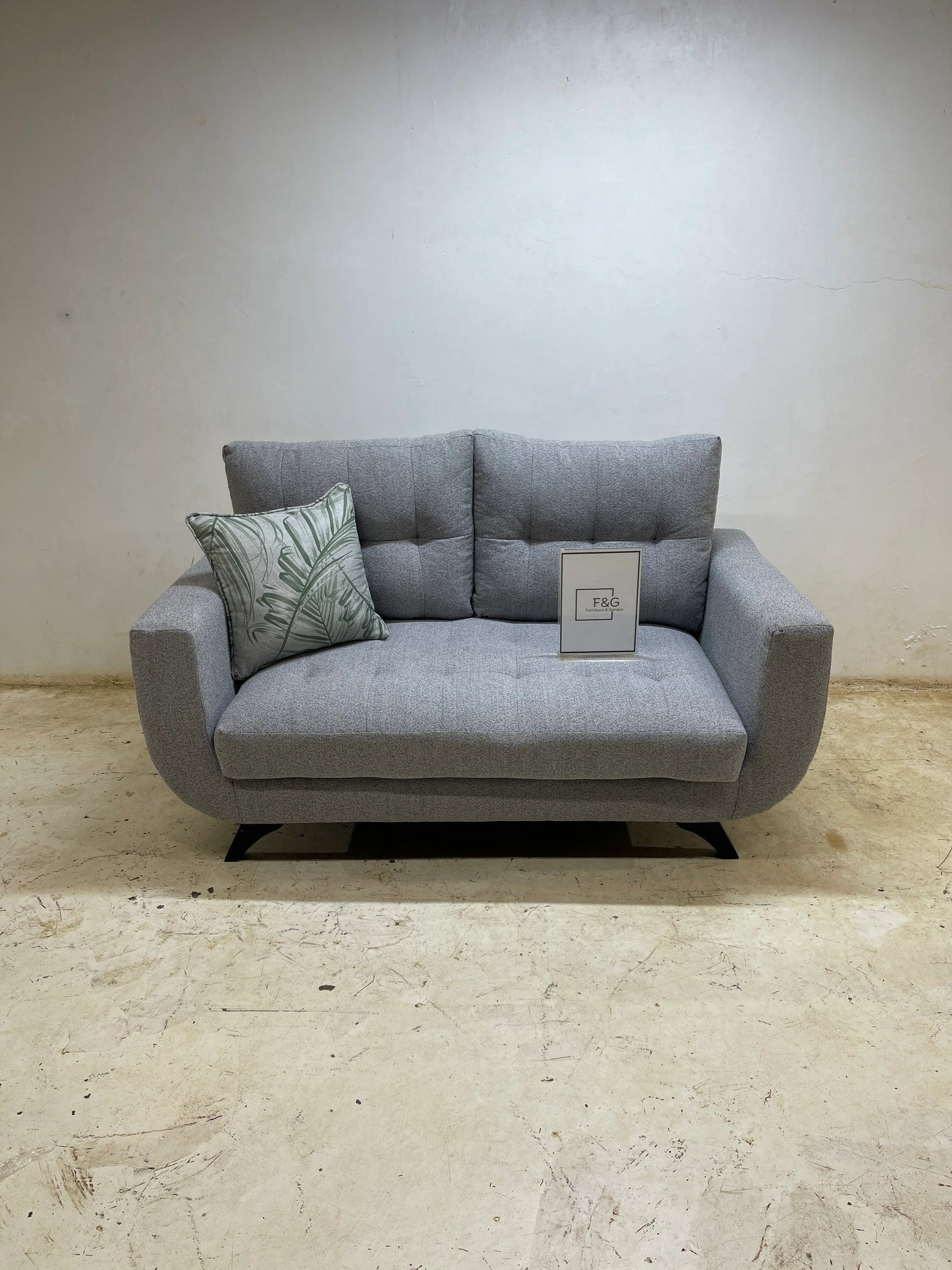 Modern grey fabric loveseat sofa with tufted cushions and decorative leaf print pillow on concrete floor