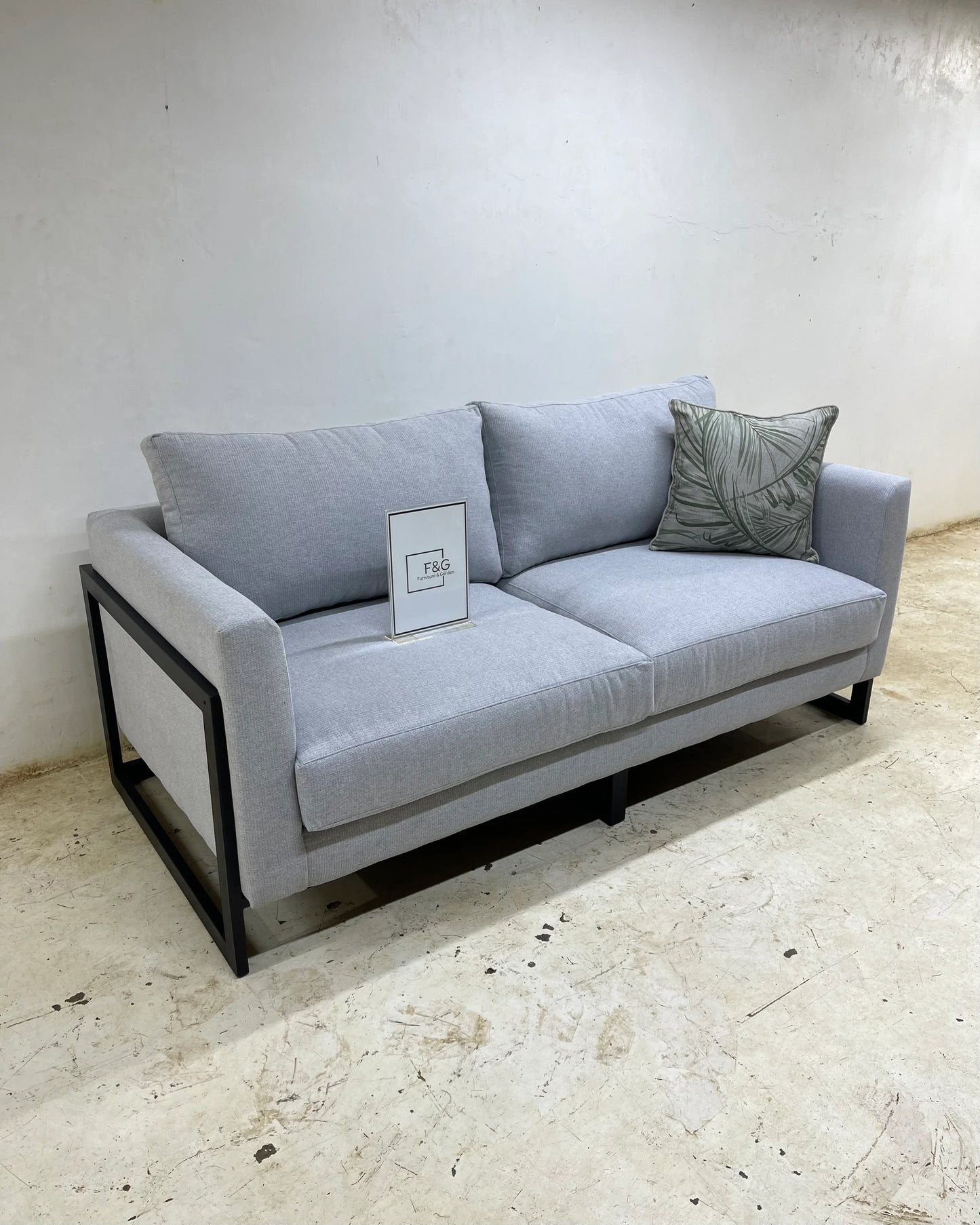 Modern light gray fabric sofa with black metal frame, decorative leaf-patterned pillow, and F&G sign