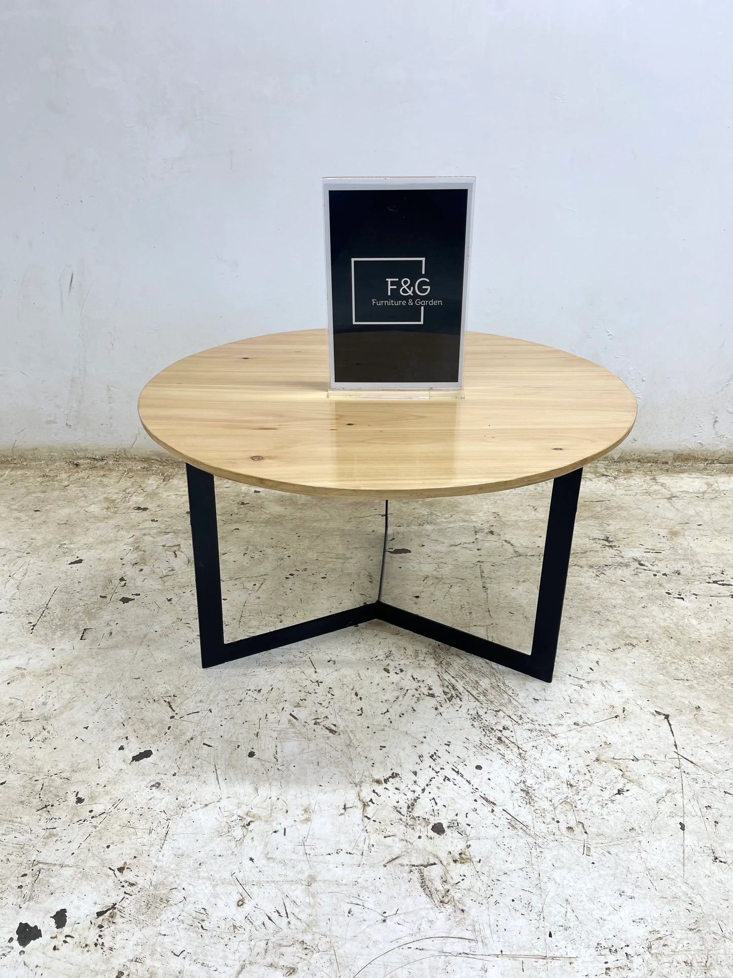 Round wooden coffee table with black metal legs on worn concrete floor, F&G Furniture & Garden sign