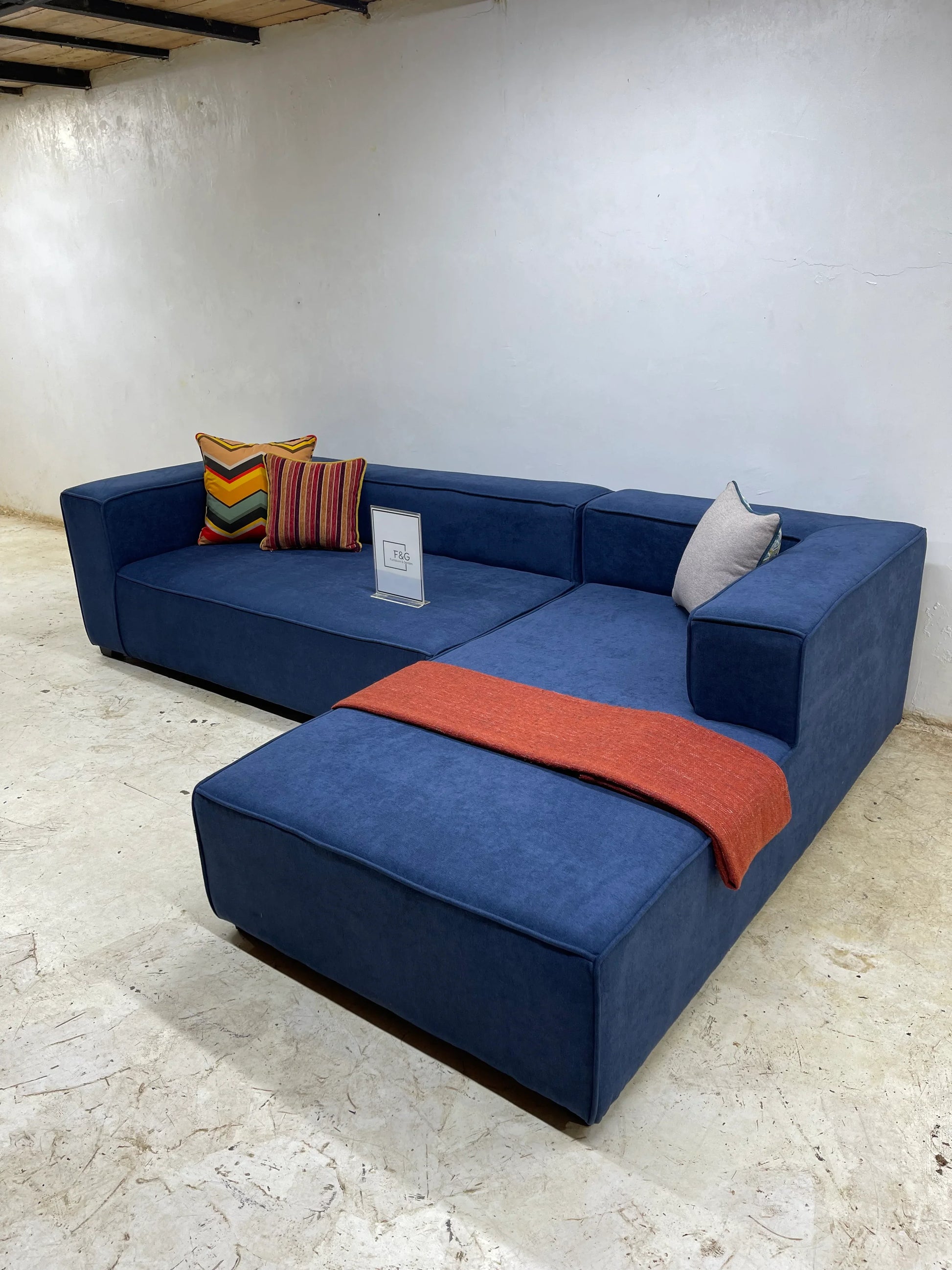 Blue sectional sofa with multicolored and gray cushions and an orange throw in a minimalist room